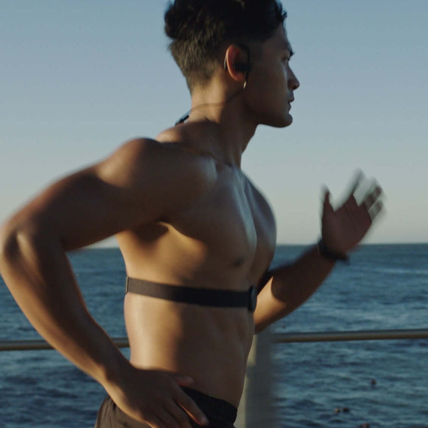 A man running by the ocean while wearing a heart monitor strap on his chest.