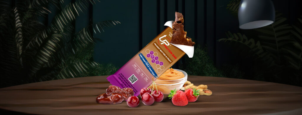 A picture of a protein bar by Up Proteins, on a table with fruit around it, with a wrapper that says peanut butter and fruit, and part of the bar sticking out.