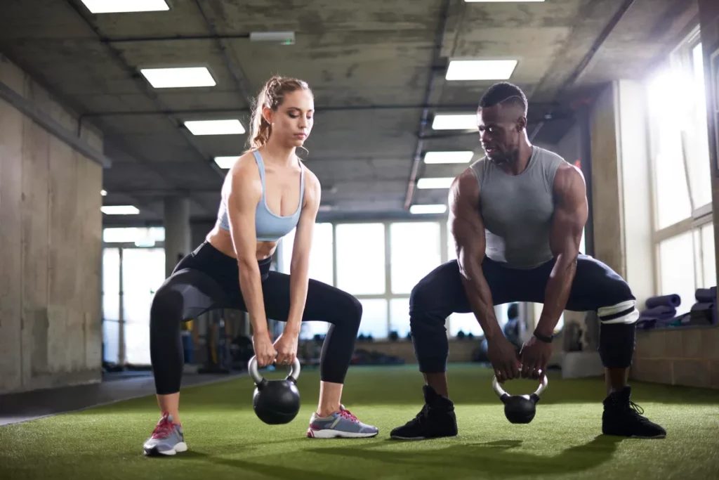 A picture of a man and woman in a gym, each doing squats while holding a kettlebell between their legs.