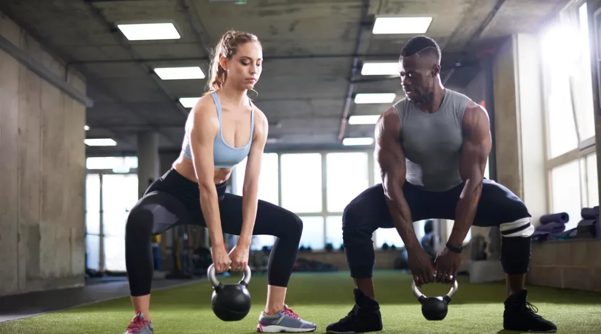 A picture of a man and woman in a gym, each doing squats while holding a kettlebell between their legs.