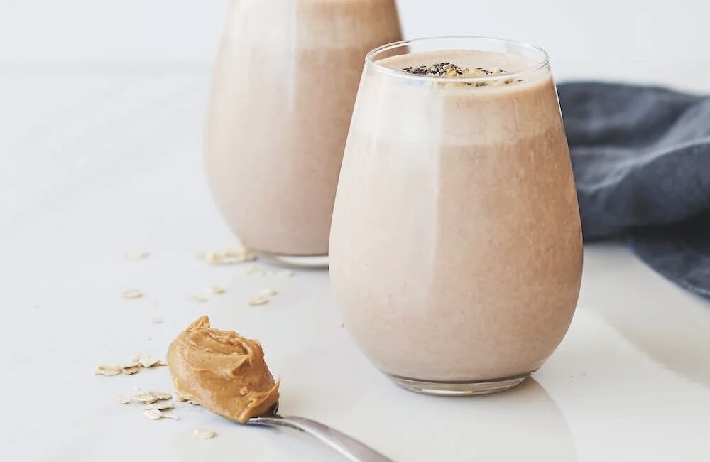 Image of two light brown colored smoothies, in jars, with a spoon full of peanut butter sitting on the counter nearby