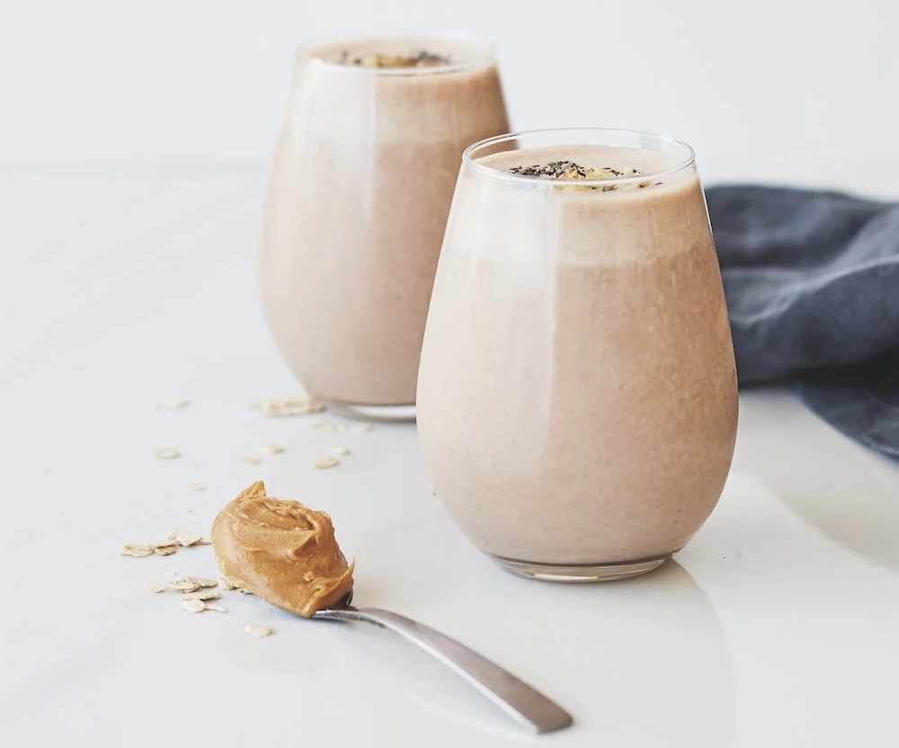 Image of two light brown colored smoothies, in jars, with a spoon full of peanut butter sitting on the counter nearby