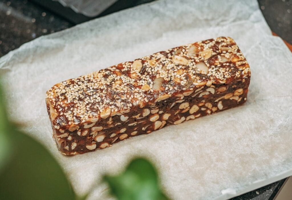 A brown protein bar, with light colored seeds and nuts in it, sitting on paper wrap