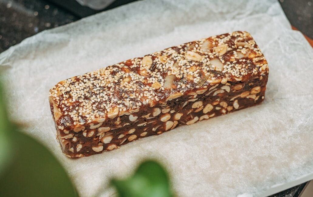 A brown protein bar, with light colored seeds and nuts in it, sitting on paper wrap