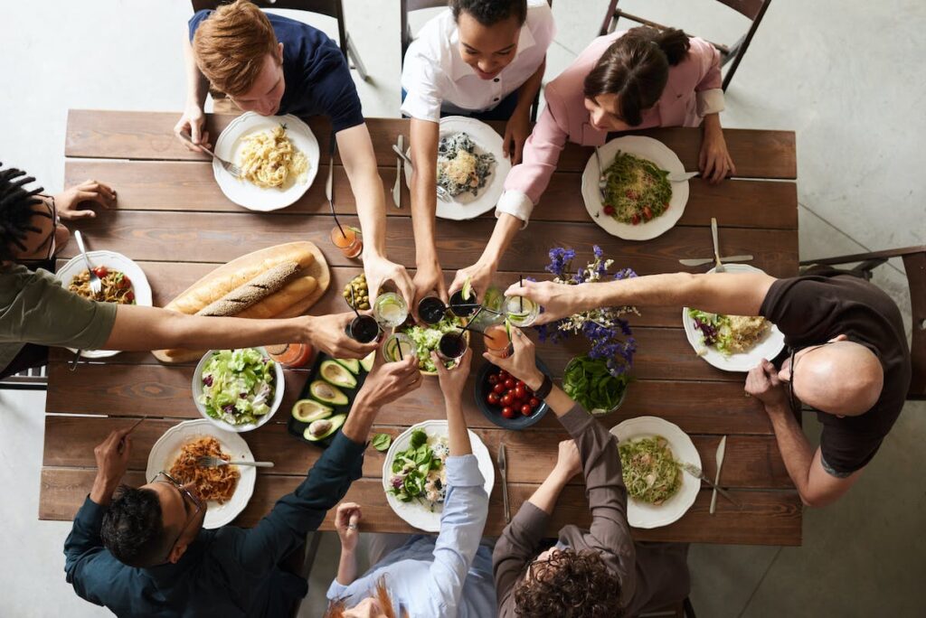 A picture taken from above, of 6 people sitting at a table with food on it, putting their glasses to the center of the table for a toast.