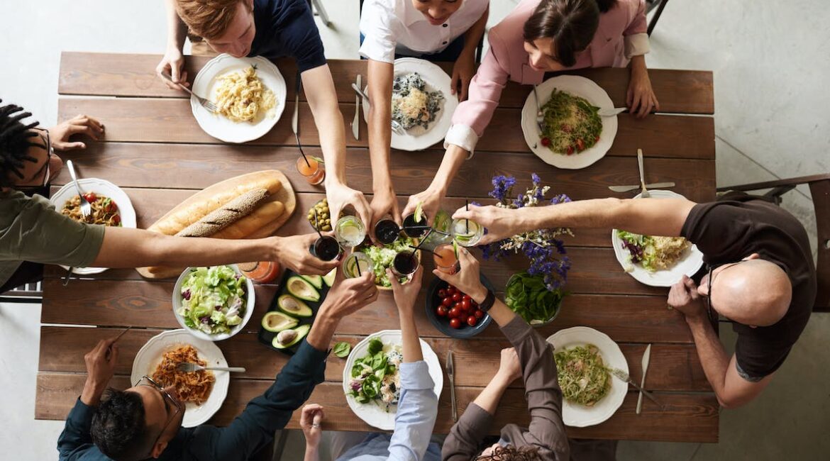 A picture taken from above, of 6 people sitting at a table with food on it, putting their glasses to the center of the table for a toast.