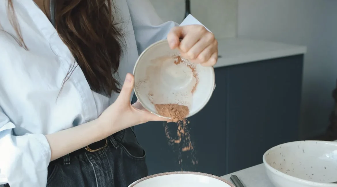 A picture of a woman pouring a brown powder into a bowl with white flour.