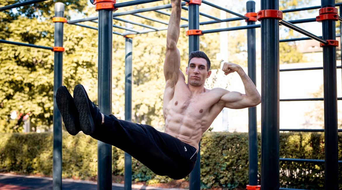 A picture of a muscular man hanging from a bar by one arm while flexing the other.