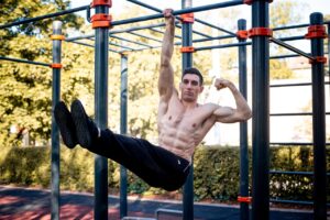 A picture of a muscular man hanging from a bar by one arm while flexing the other.
