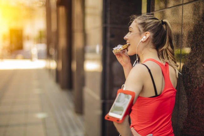 A woman outside, wearing running clothing, eating a protein bar.